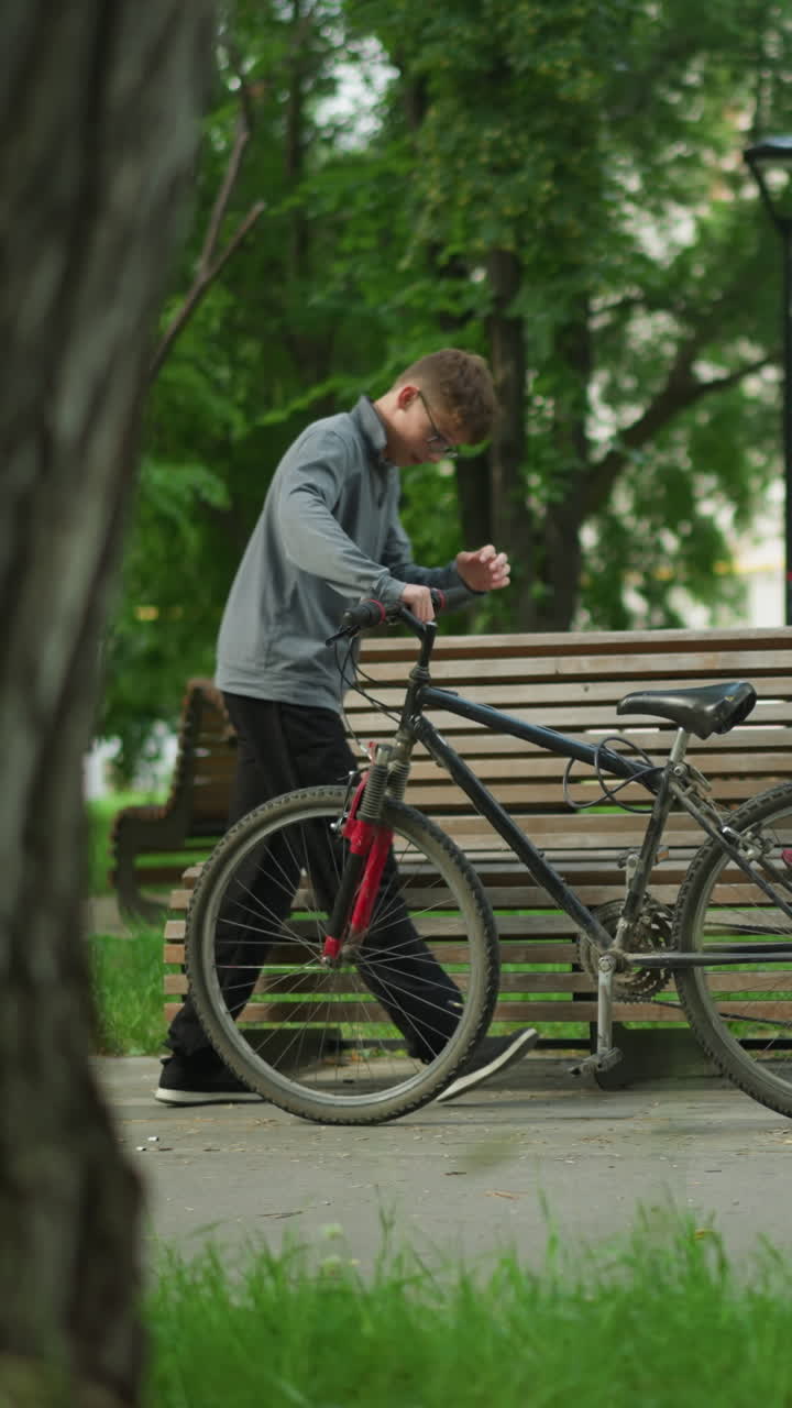 어린 소년은 공원으로 자전거를 타고 나무 벤치에 앉아 자전거의 바를 만져서 자세히 조사합니다. 초록색 나무, 풀, 그리고 배경의 주거 건물로 둘러싸여 있습니다.