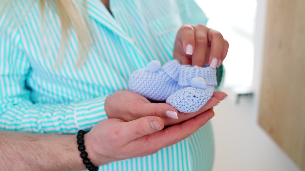 Unrecognized pregnant lady holding tiny blue socks for future kid. Male hand is supporting from below. Close up.