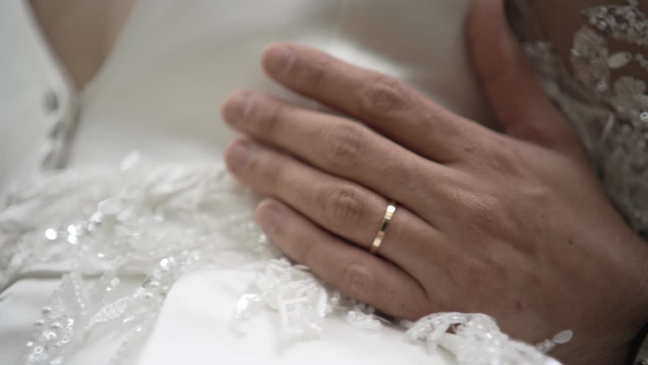 Close up of a groom's hand with wedding ring resting on a lace-decorated bridal gown