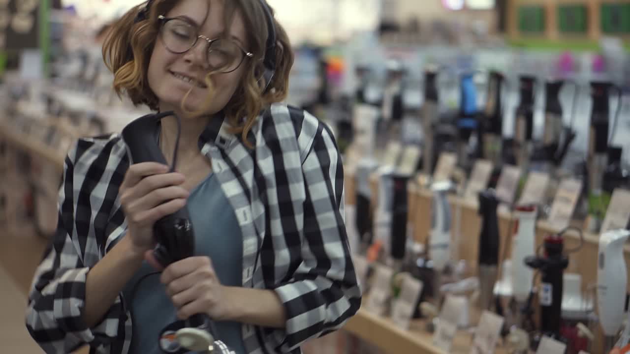 Portrait of cute caucasian pretty young woman with short curly hair, plaid shirt and in headphones dancing and pretending she's singing in the middle of the supermarket with household using blender stick as microphone