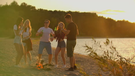 company of young people are dancing in shorts and t-shirts around bonfire on the open air party with beer. they are enjoying the summer evening at sunset near the forest.