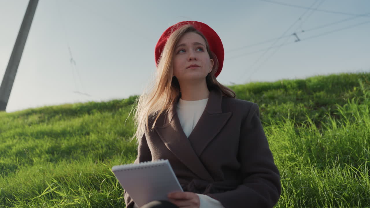 Giovane donna bianca che disegna su un blocco note in un parco erboso illuminato dal sole, indossa un basco rosso e un cappotto. Capelli mossi dal vento, zaino al suo fianco, espressione calma e pensierosa, atmosfera studentesca casual, calda luce dorata.