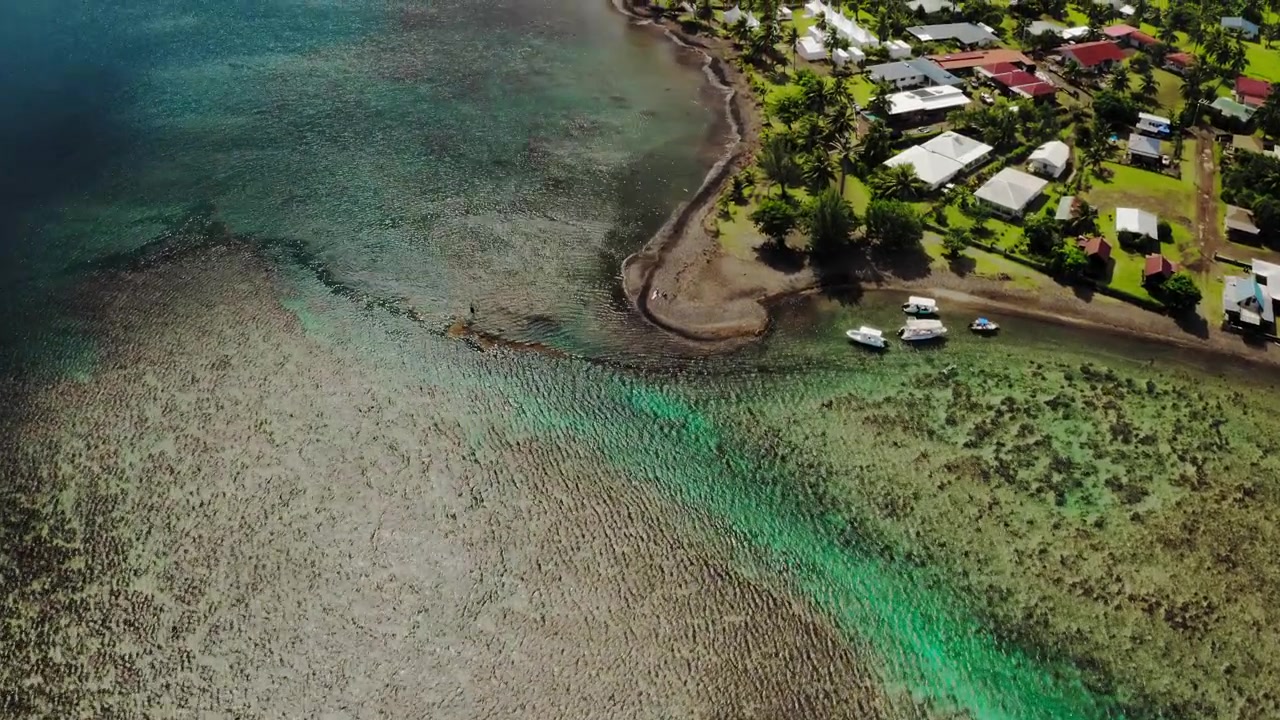 Ripresa con drone dall'alto su motivi di barriera corallina testurizzata vicino a Teahupoo, Tahiti, con il bordo di un villaggio costiero visibile all'angolo dell'inquadratura