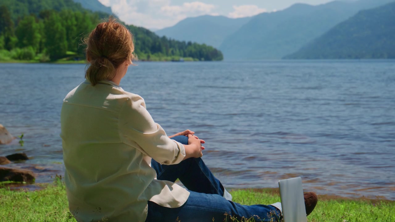 Eine Frau sitzt friedlich an einem ruhigen See und blickt auf ferne Berge. Sie wirkt entspannt und genießt die stille Einsamkeit im Freien. Diese Szene ist ideal für Reise-, Meditations- oder Naturprojekte.