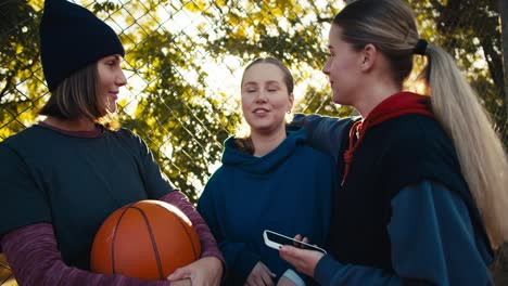 happy three blonde girls in sportswear communicate and hold an orange basketball ball on a summer street playground near green trees on a summer sunny day