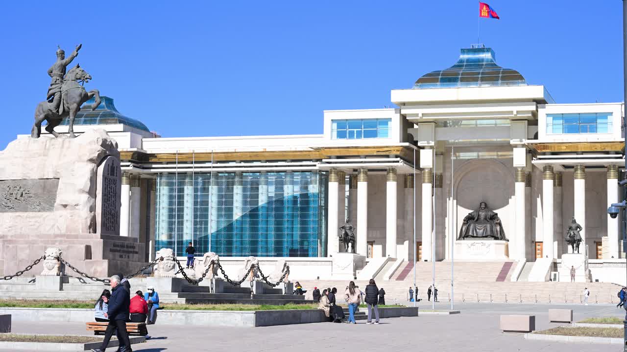 On a sunny day, visitors at Sukhbaatar Square admire the statues of Damdin Sukhbaatar (left) and Genghis Khan, with the Government Palace and Mongolian flag in the background in Ulaanbaatar.