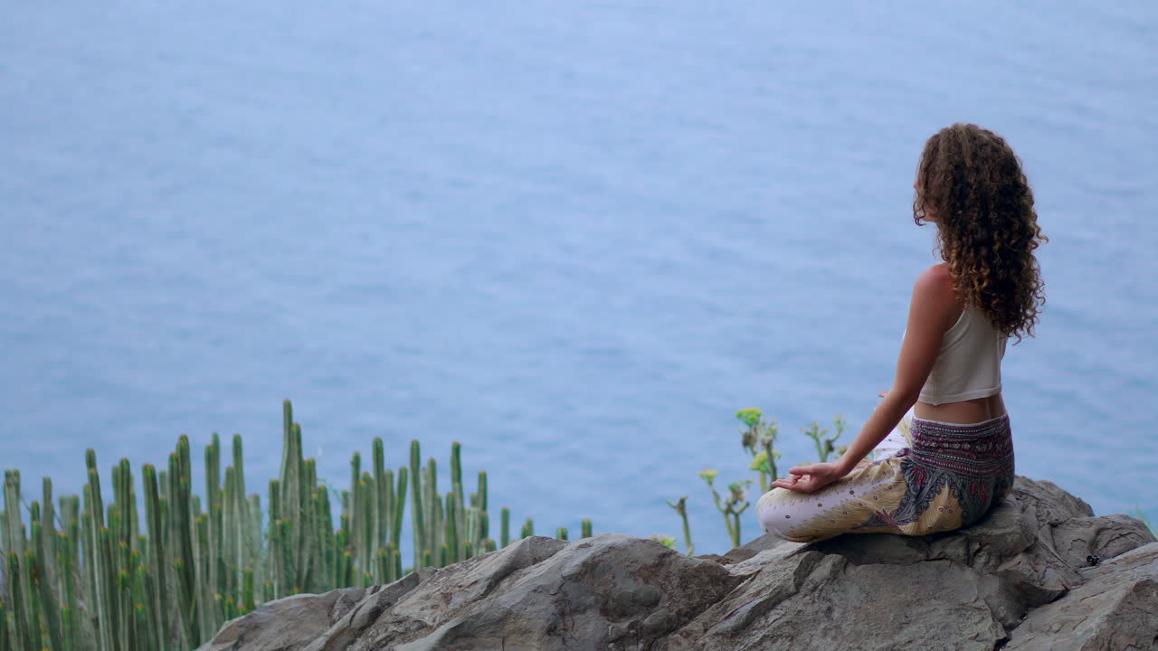 Amidst island mountains, a young woman practices yoga in Lotus position on a rock at the mountain's summit, overlooking the ocean