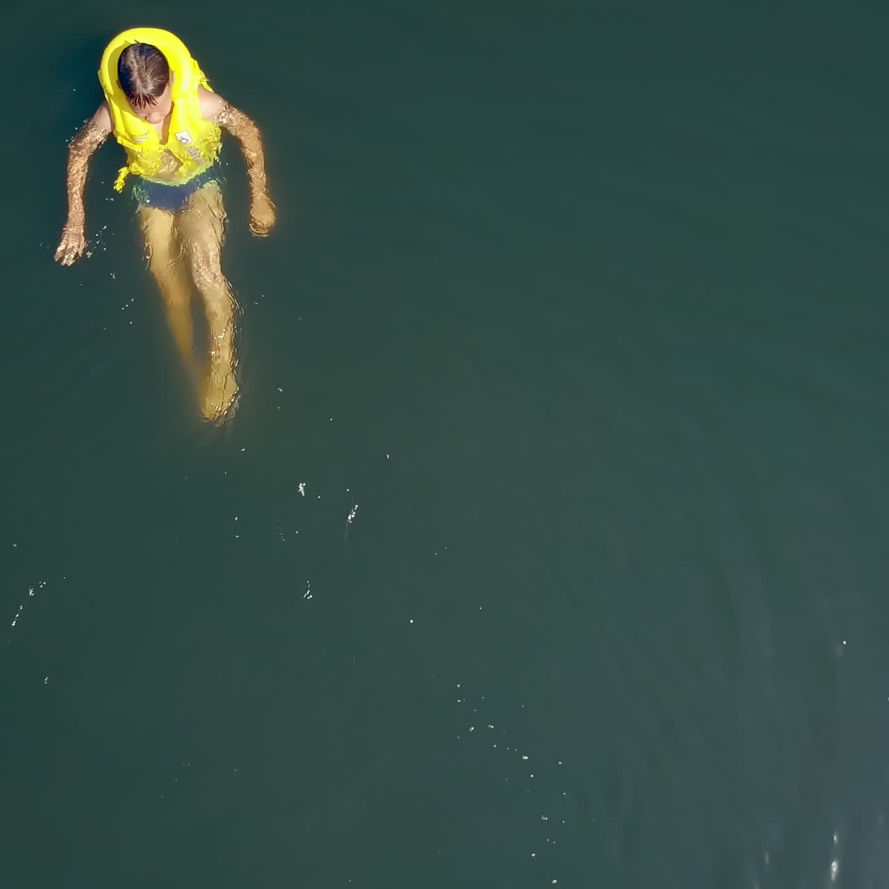 Active kid in life yellow waistcoat swimming alone in the clear water in bright summer day. Portrait of cute funny boy bathing on his back in water swinging legs. Motion camera bottom up