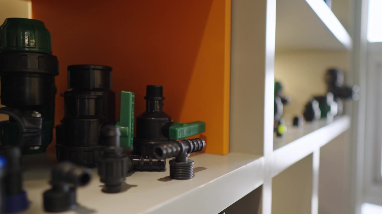 Close-up shot with rack focus movement across a warehouse shelf displaying various black and green plastic plumbing fittings, valves, and irrigation connectors for industrial water systems