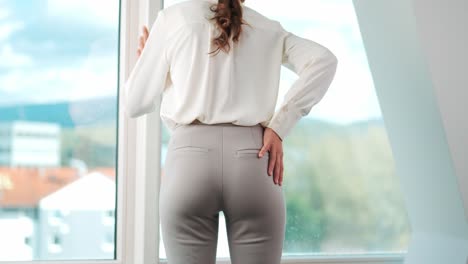 back view of young woman with back pain in front of window in modern apartment office, back problems, incorrect posture