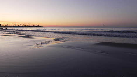 Die-Skyline-Der-Morgendämmerung-Spiegelt-Sich-Am-Strand,-Während-Ein-Mann-über-Die-Küste-Geht