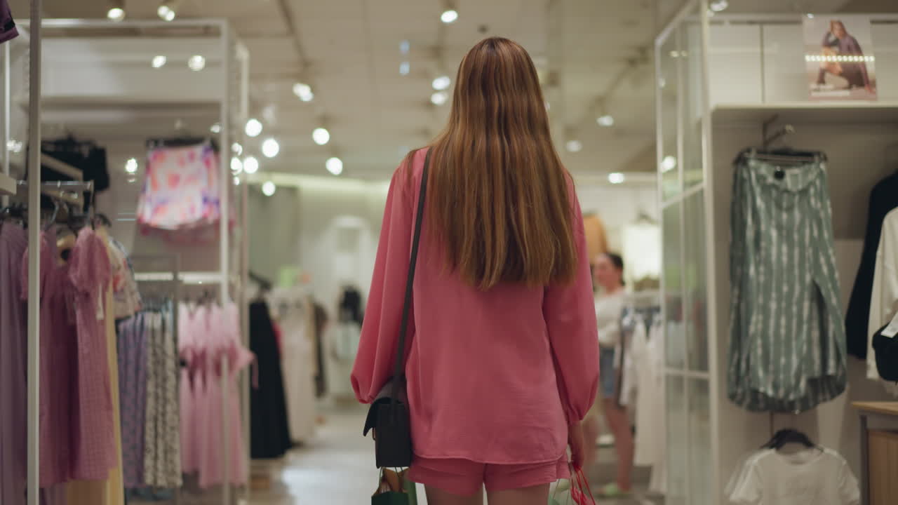 백인 여성이 두 개의 쇼핑 가방을 들고 선반과 선반에 끔하게 배열된 옷을 가진 옷 가게를 걸어가는 뒷면, 배경에서 지나가는 두 명의 여성의 약간 흐릿한 시각
