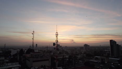 En-Primer-Plano-Se-Ve-Una-Antena-De-Telecomunicaciones-En-El-Fondo-Se-Ven-Volcanes-Durante-Un-Amanecer-En-Una-Ciudad