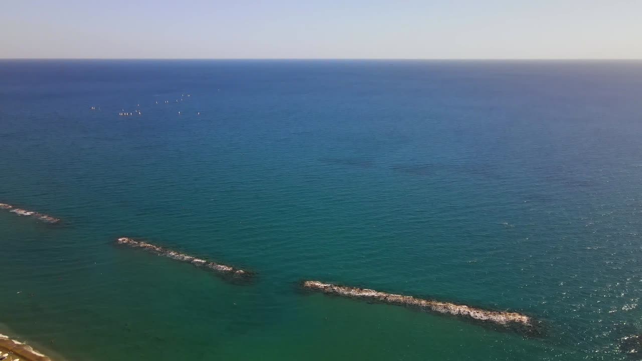 Drone video capturing tranquil Durrës coastline with breakwaters and scattered buoys under clear skies and serene blue waters, offering a peaceful seascape from above