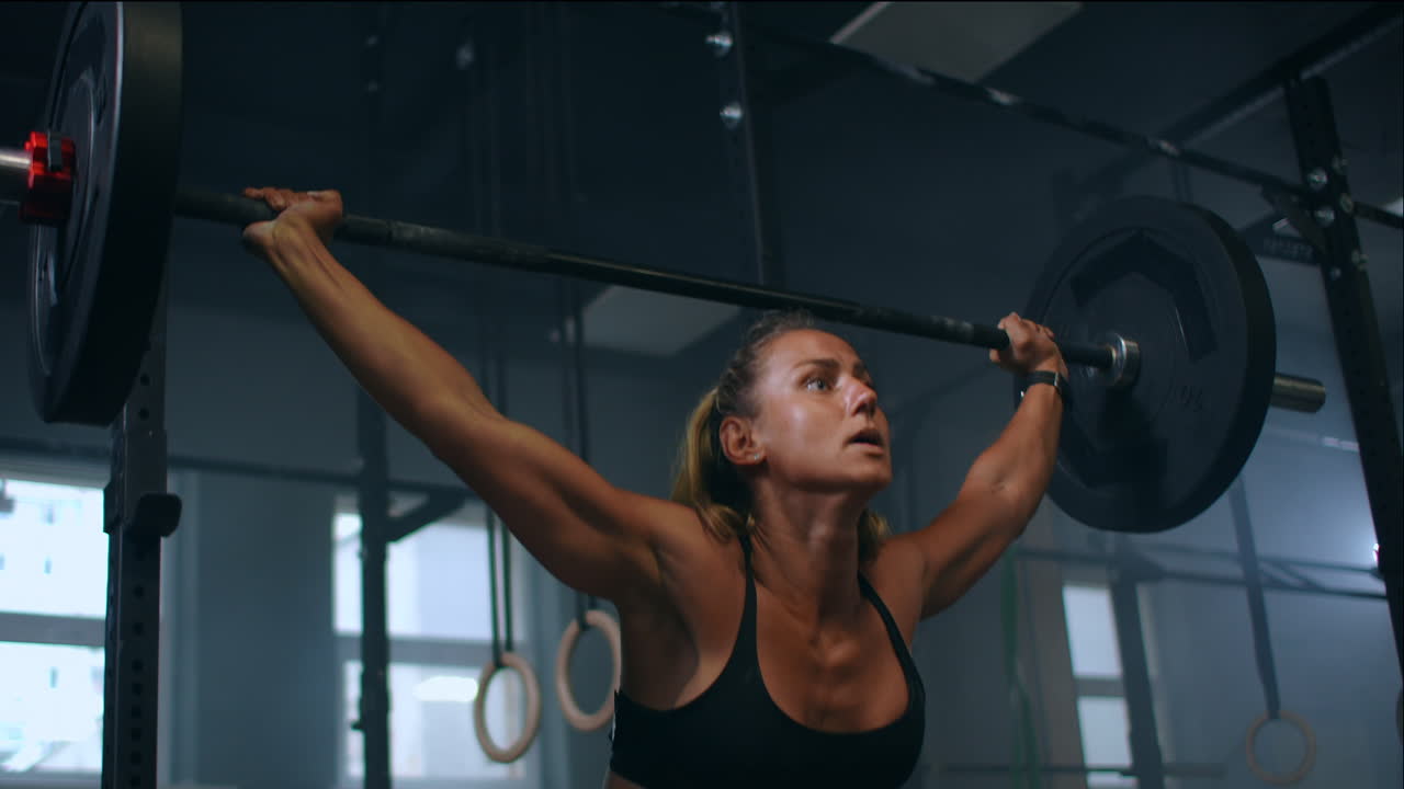 Ralenti : entraînement de squats avec barre au-dessus de la tête. Femme musclée soulevant une barre au-dessus de la tête pour entraîner ses épaules à la salle de sport. Femme de fitness confiante pratiquant un entraînement de force avec équipement lourd.