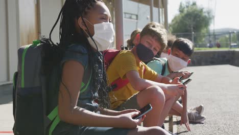 Group-of-kids-wearing-face-masks-using-smartphones-while-sitting-together