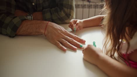 Close-up-shot-of-a-little-brunette-girl-in-a-pink-dress-doing-a-manicure-to-her-dad,-a-man-in-a-green-checkered-shirt,-using-green-polish-on-the-kitchen-table-at-home