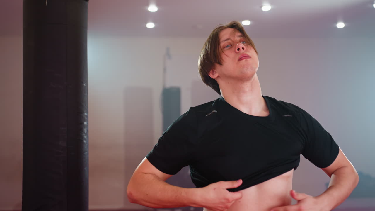 Fighter put on a black shirt in gym environment, standing near punching bag under strong lights, showing chest and torso while preparing for workout, representing strength