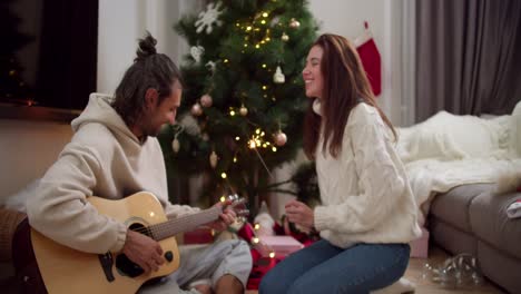 feliz chico moreno en un suéter blanco toca la guitarra y su novia morena canta y sostiene chispas en sus manos cerca del árbol de año nuevo y regalos en una acogedora habitación de invierno