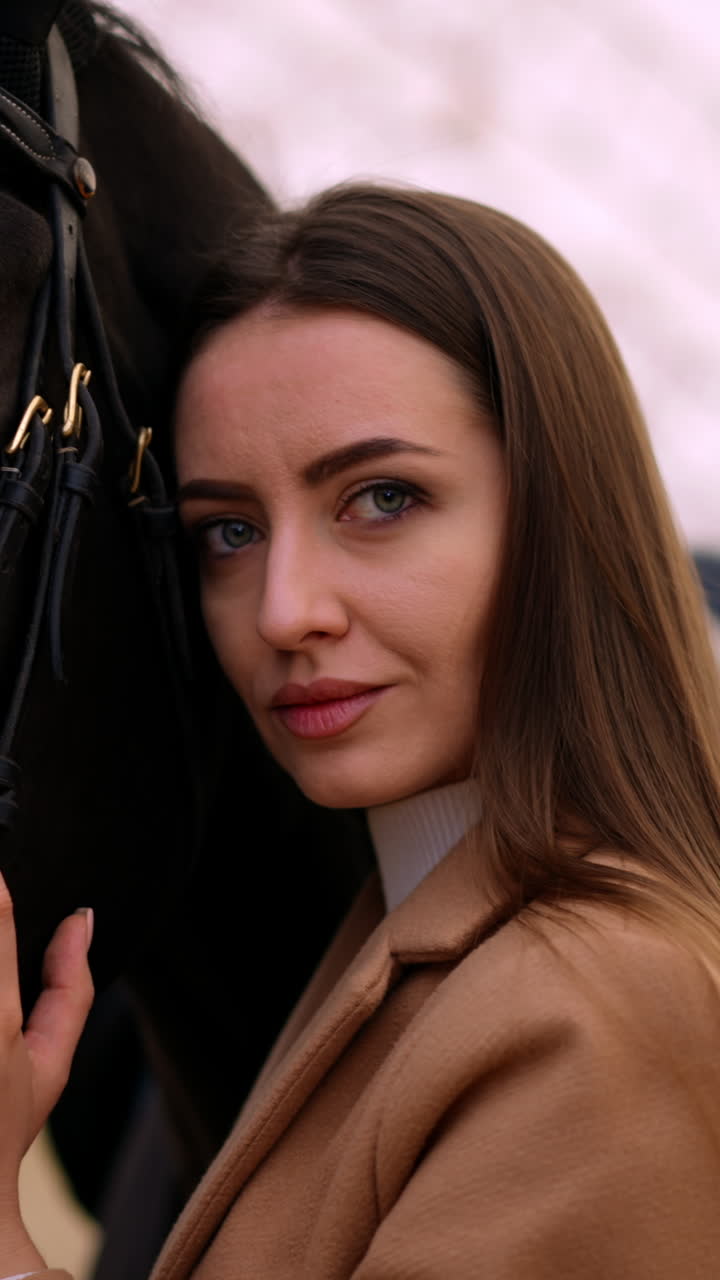 Beautiful big-eyed Caucasian woman leaned to a graceful black horse. Close up. Riding lessons at drill hall. Blurred backdrop. Vertical video
