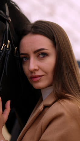 beautiful big-eyed caucasian woman leaned to a graceful black horse. close up. riding lessons at drill hall. blurred backdrop. vertical video