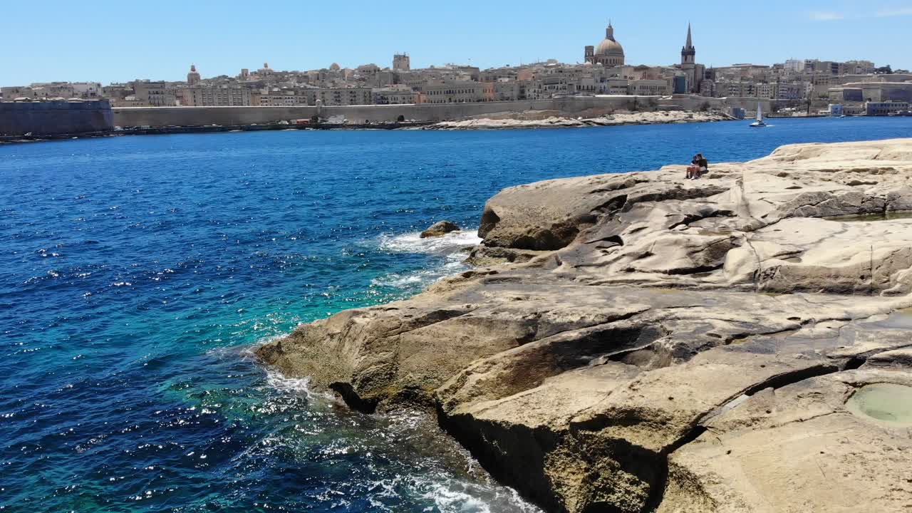 Veduta aerea verso La Valletta, Malta, con onde che si infrangono sulla rocciosa spiaggia calcarea di Sliema in una soleggiata giornata estiva.