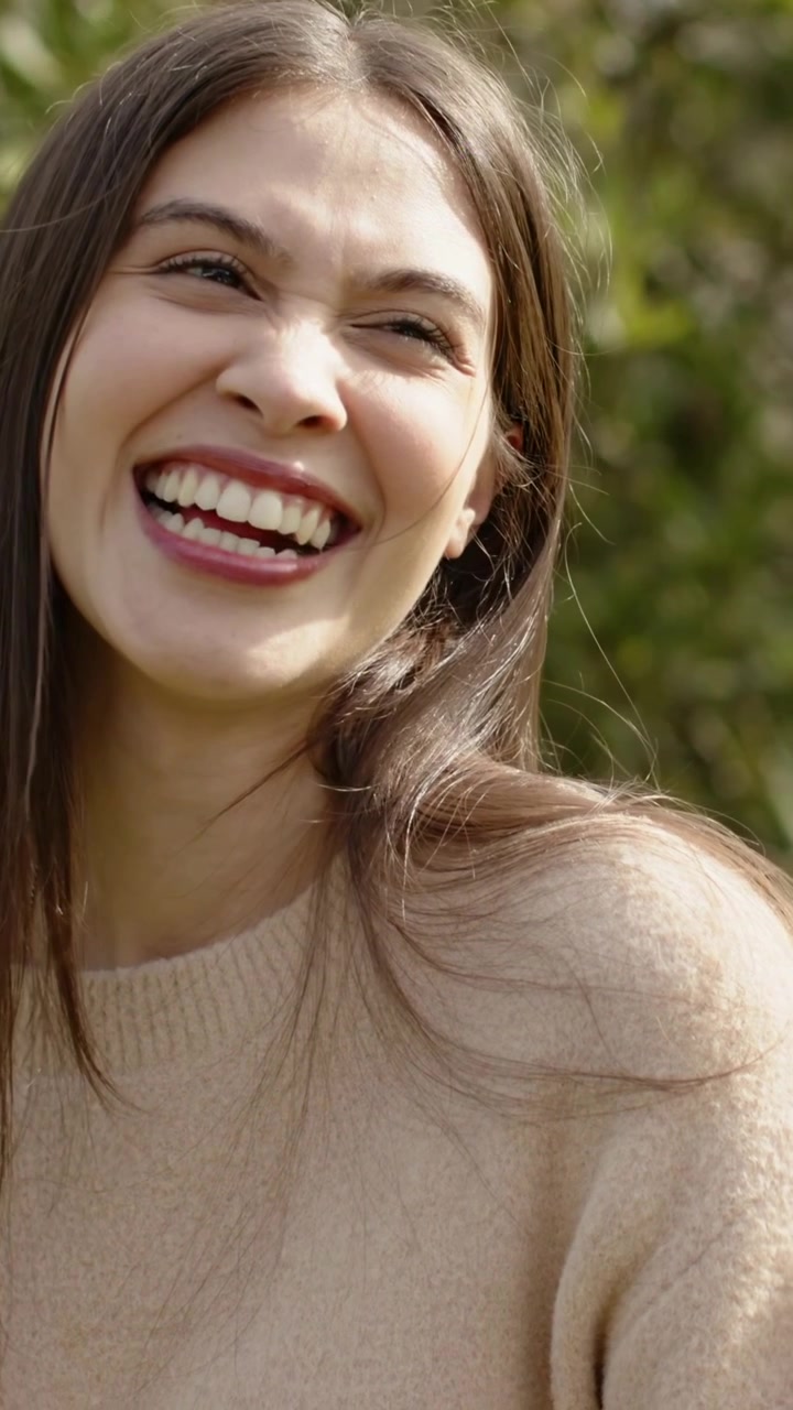 Video vertical: Mujer sonriendo y riendo después de una señal fuera de cámara, moviéndose hacia la izquierda en el parque vistiendo un suéter de punto beige.