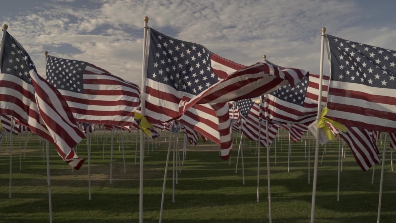 Rows of American flags decorated with yellow ribbons wave heavily in the wind at an outdoor memorial park near sunset. The static wide shot captures the patriotic display of honor and remembrance.