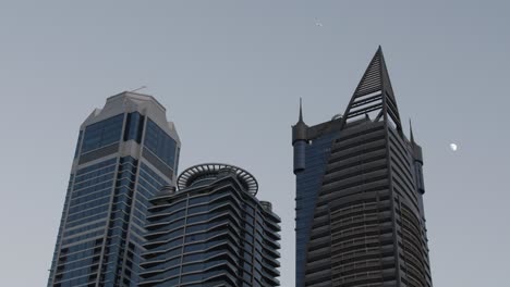 a distant airplane passes by behind the tops of three dubai skyscrapers with the moon visible in the distance