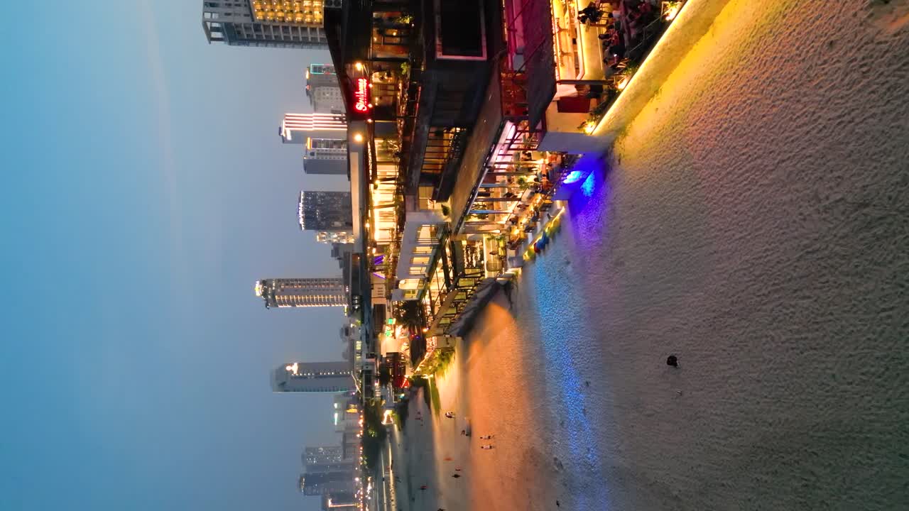 Illuminated beachfront restaurants and coracle boats on the sand at sunset and the city skyline in the background in Da Nang, Vietnam, drone revealing, vertical shot
