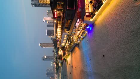 illuminated beachfront restaurants and coracle boats on the sand at sunset and the city skyline in the background in da nang, vietnam, drone revealing, vertical shot
