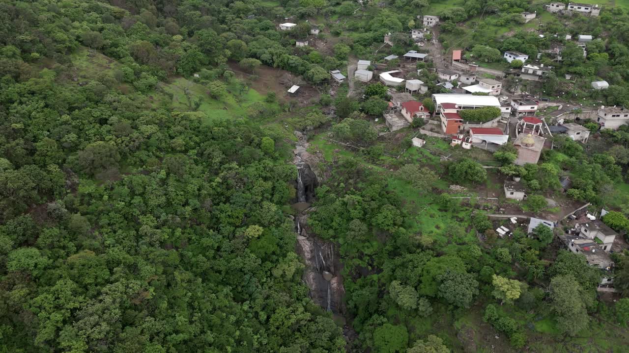 Vista aerea con drone di un piccolo villaggio accanto a una fitta foresta con una stretta cascata che scorre attraverso la valle circondata dal verde in un paesaggio naturale sotto una luce diurna soffusa.
