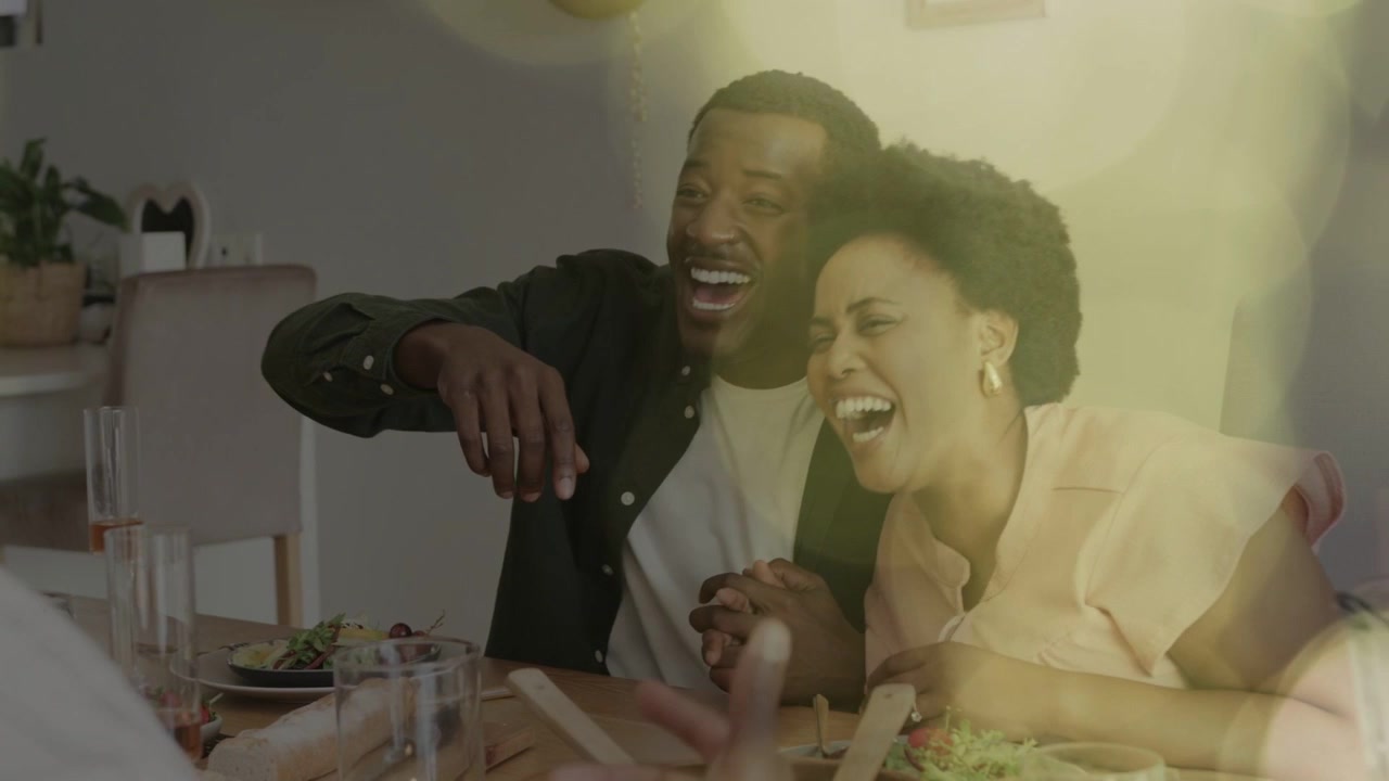 Casal reagindo ao movimento de convidado à mesa, rindo e se inclinando, com bokeh os cercando e destacando a comida.