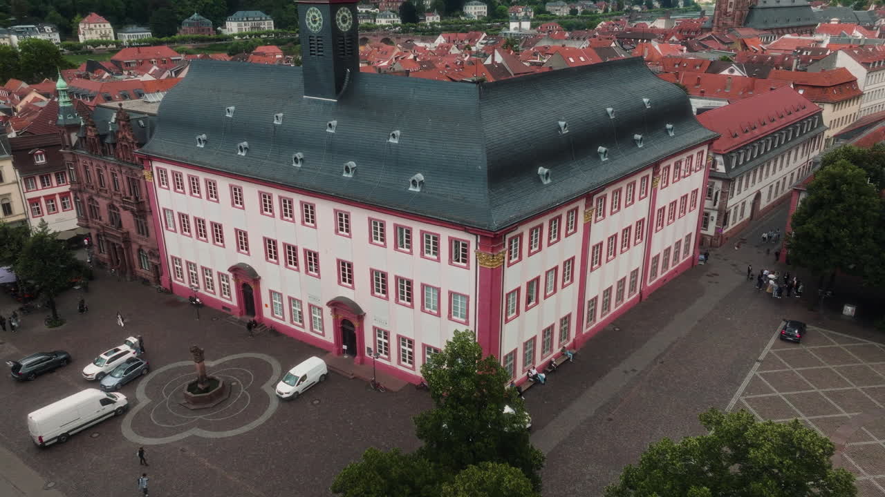 Drone pulling back and rising above a red and white historic building, revealing surrounding rooftops, streets, and the river