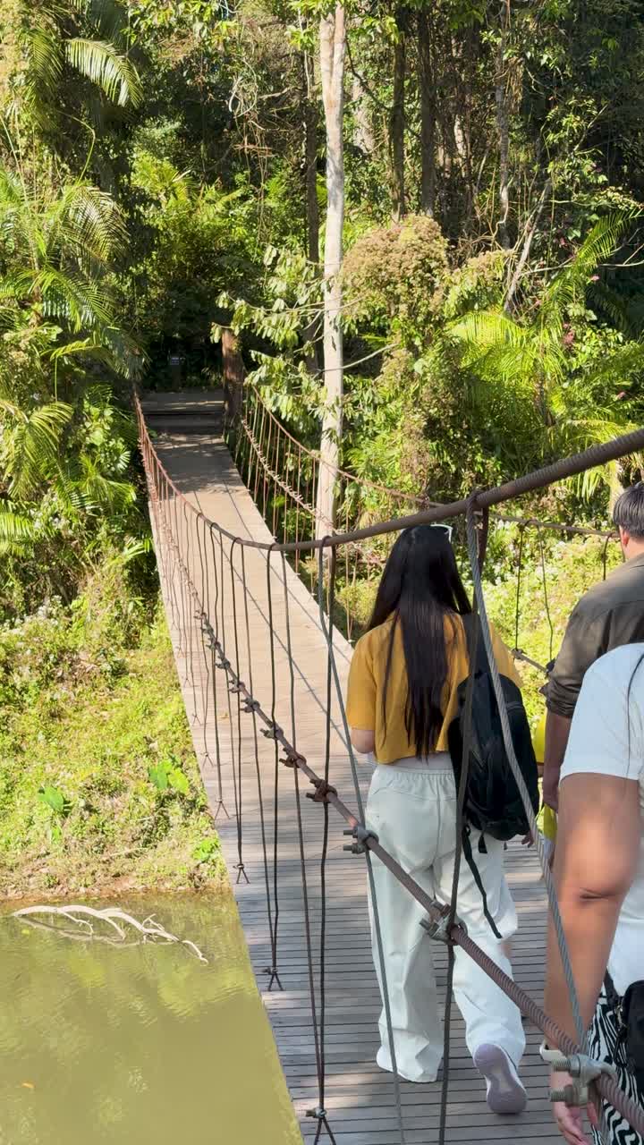 Wisatawan menyeberangi jembatan gantung kayu di atas sungai di tengah hutan tropis yang rimbun.