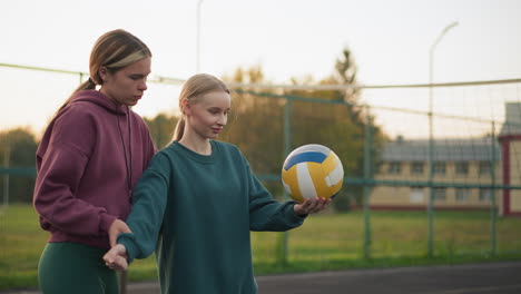 volleyball instructor demonstrating serve technique to beginner, then letting her serve ball, both standing on outdoor court with volleyball in hand, distant football field in background