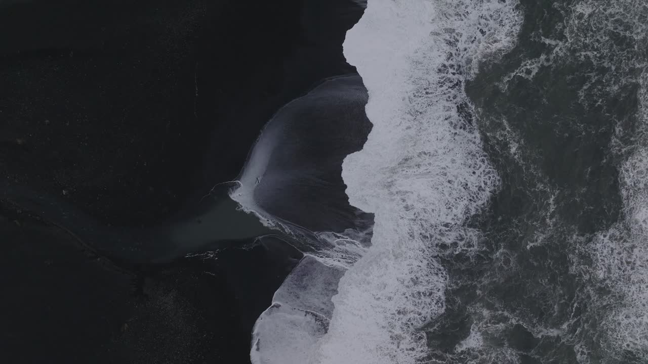 Aerial top view of ocean waves crashing on Iceland S&oacute;lheimasandur black sand beach, on a moody day