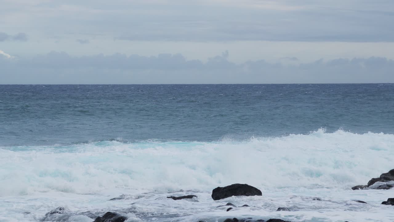 Wide shot of rough ocean waves colliding with dark coastal rocks, foamy white surf rising high against the backdrop of a cloudy horizon