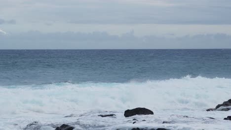 wide shot of rough ocean waves colliding with dark coastal rocks, foamy white surf rising high against the backdrop of a cloudy horizon