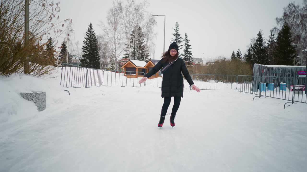 Lady skating on outdoor ice rink in winter wearing black coat, knit hat, scarf, red skates, patterned gloves, surrounded by snow, pine trees, metal fences , enjoying seasonal fitness activity