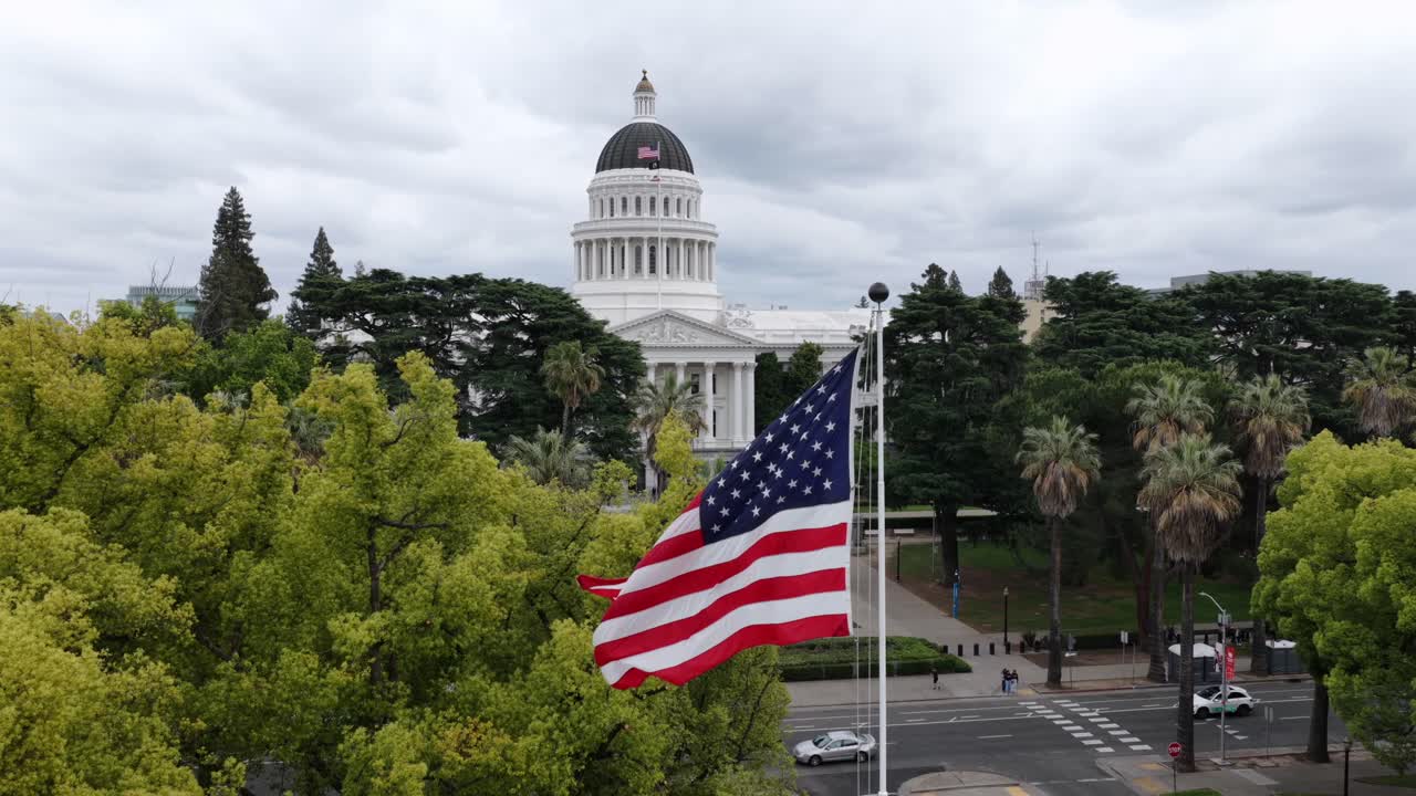 Vista aerea con drone della bandiera degli Stati Uniti che sventola davanti al Campidoglio dello Stato della California, circondato da alberi e architettura civica a Sacramento, California, USA