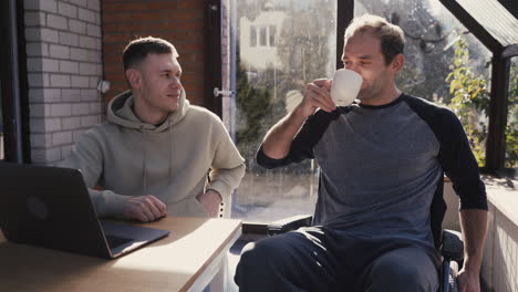 disabled man in wheelchair drinking from a coffee mug and talking with his friend at home