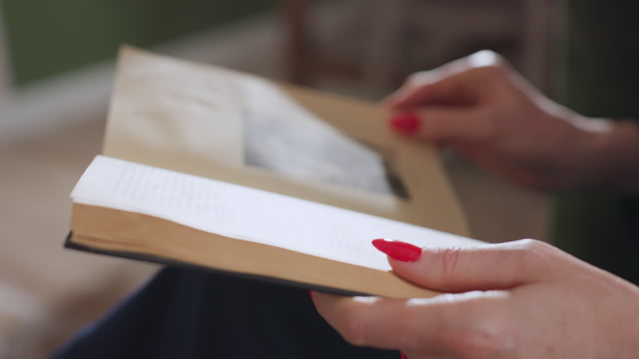 Close up of female hands with red nails gently holding open book on lap, pages spread as she reads in calm indoor setting, sunlight casting soft shadows and enhancing atmosphere of quiet focus