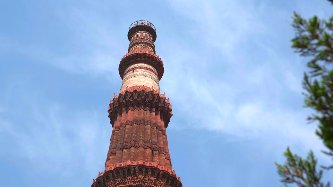 A static low-angle shot of the Qutb Minar, the world's tallest brick minaret, showcasing its intricate red sandstone carvings and marble sections against a clear blue sky in New Delhi.