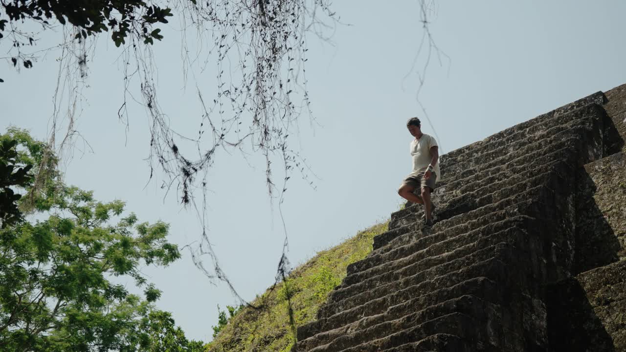 A man descends carefully from a steep Mayan pyramid at Tikal, Guatemala, framed by hanging vines and dense jungle in the background.