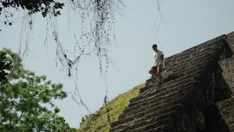 a man descends carefully from a steep mayan pyramid at tikal, guatemala, framed by hanging vines and dense jungle in the background.
