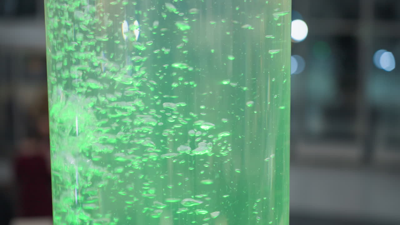 Close-up of large cylindrical water feature with green glowing water and floating bubbles, soft blur background and vibrant light effects create a tranquil, aesthetic atmosphere