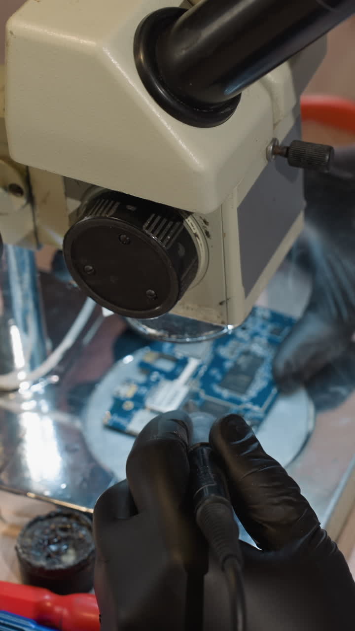 vista hacia abajo de un técnico soldando una placa de circuito bajo un microscopio, el espacio de trabajo incluye varias herramientas y equipos