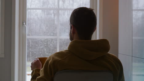 Contemplar-Al-Hombre-En-Soledad-Bebe-Bebida-Caliente-Mientras-Ve-Caer-La-Primera-Nieve-De-Invierno-A-Través-De-La-Ventana,-Vista-Trasera