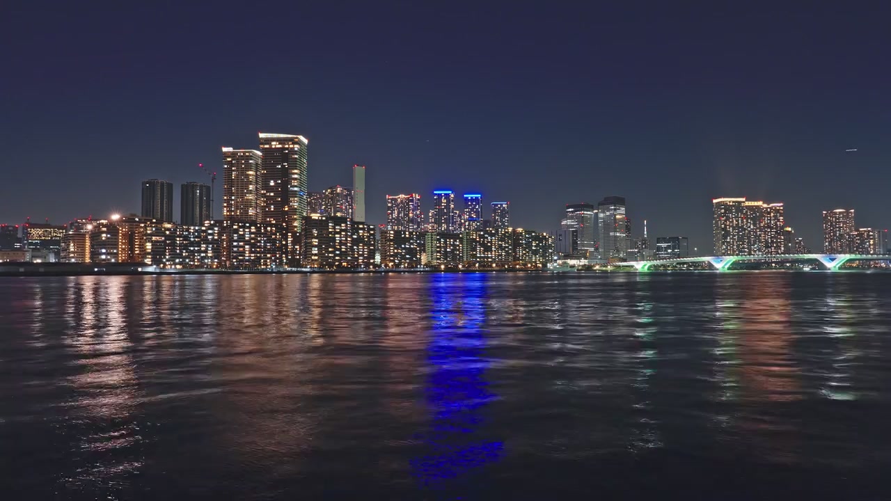 Dense residential towers glittering across the horizon, vivid blue light streak reflecting on rippling water, Harumi Bridge illumination visible right of frame at night.